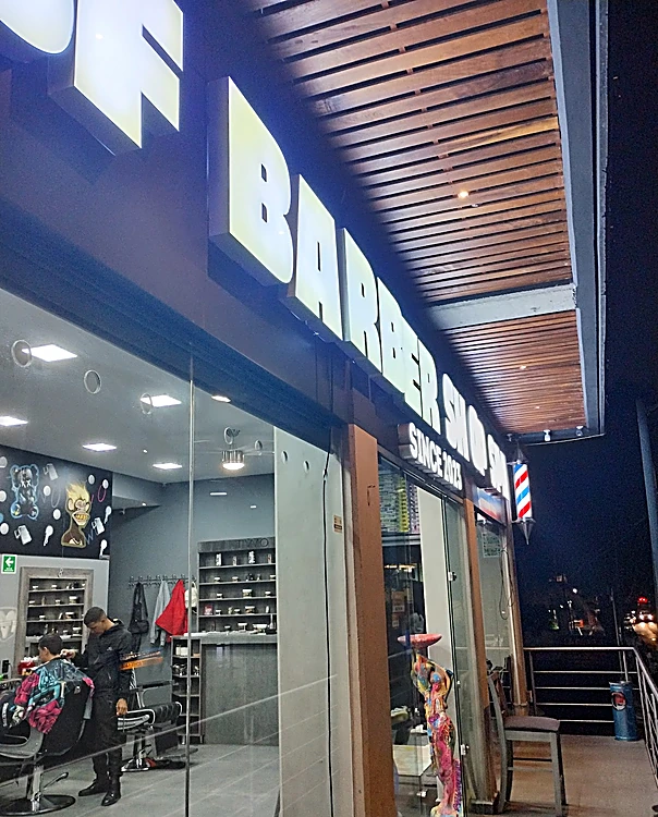 Fachada de noche: letras blancas iluminadas de 3F Barber Shop bajo un plafón de listones de madera y el poste de barbero
